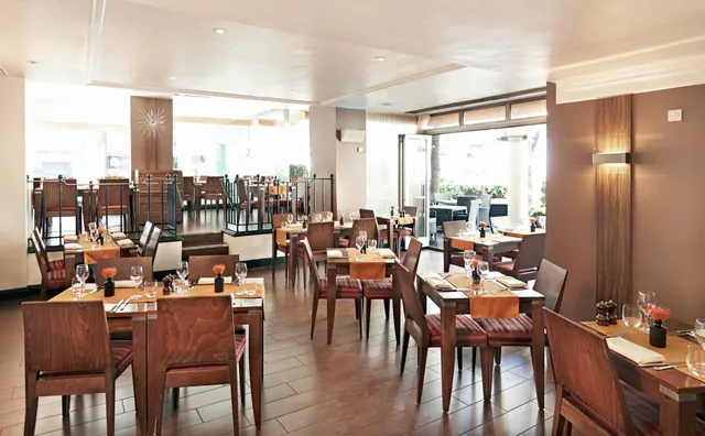 A restaurant dining room with set wooden tables, upholstered wooden chairs and large windows with greenery visible outside.