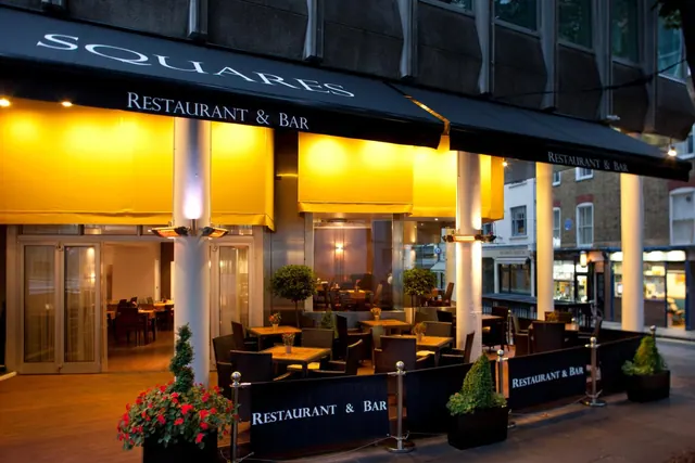 Exterior of the Squares Restaurant & Bar at dusk, with outdoor tables and chairs on a terrace under yellow awnings.