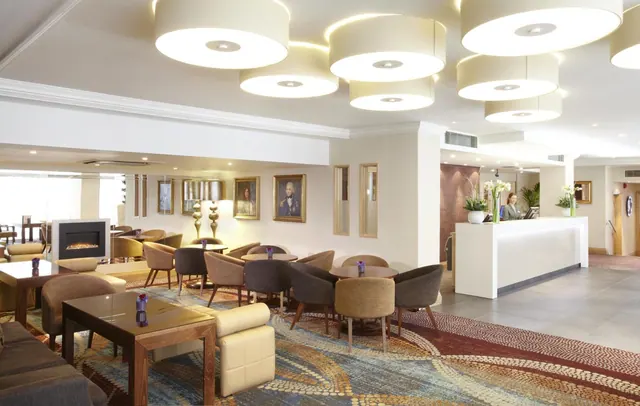 A hotel lobby with a lounge area of armchairs and small tables under large drum pendant lights, with a staff member behind the reception desk on the right.