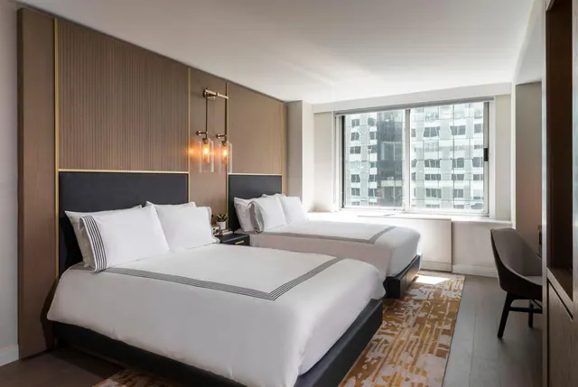 A hotel bedroom with two double beds with white bedding, a wood slatted headboard wall, a desk and chair on the right, and a window looking out onto high-rise buildings.