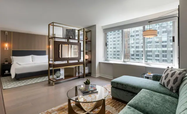 A hotel room with a king bed on the left, a green velvet sofa and round glass coffee table on the right, and large windows overlooking neighbouring high-rise buildings.
