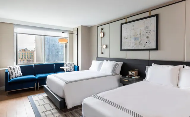 A hotel bedroom with two beds dressed in white linens, a blue velvet sofa, and a window showing city buildings outside.