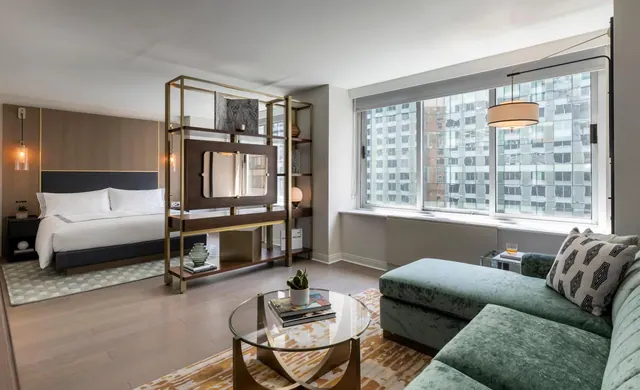 A hotel room with a king bed, a green velvet sofa, an open shelving divider, and a large window looking onto high-rise buildings.