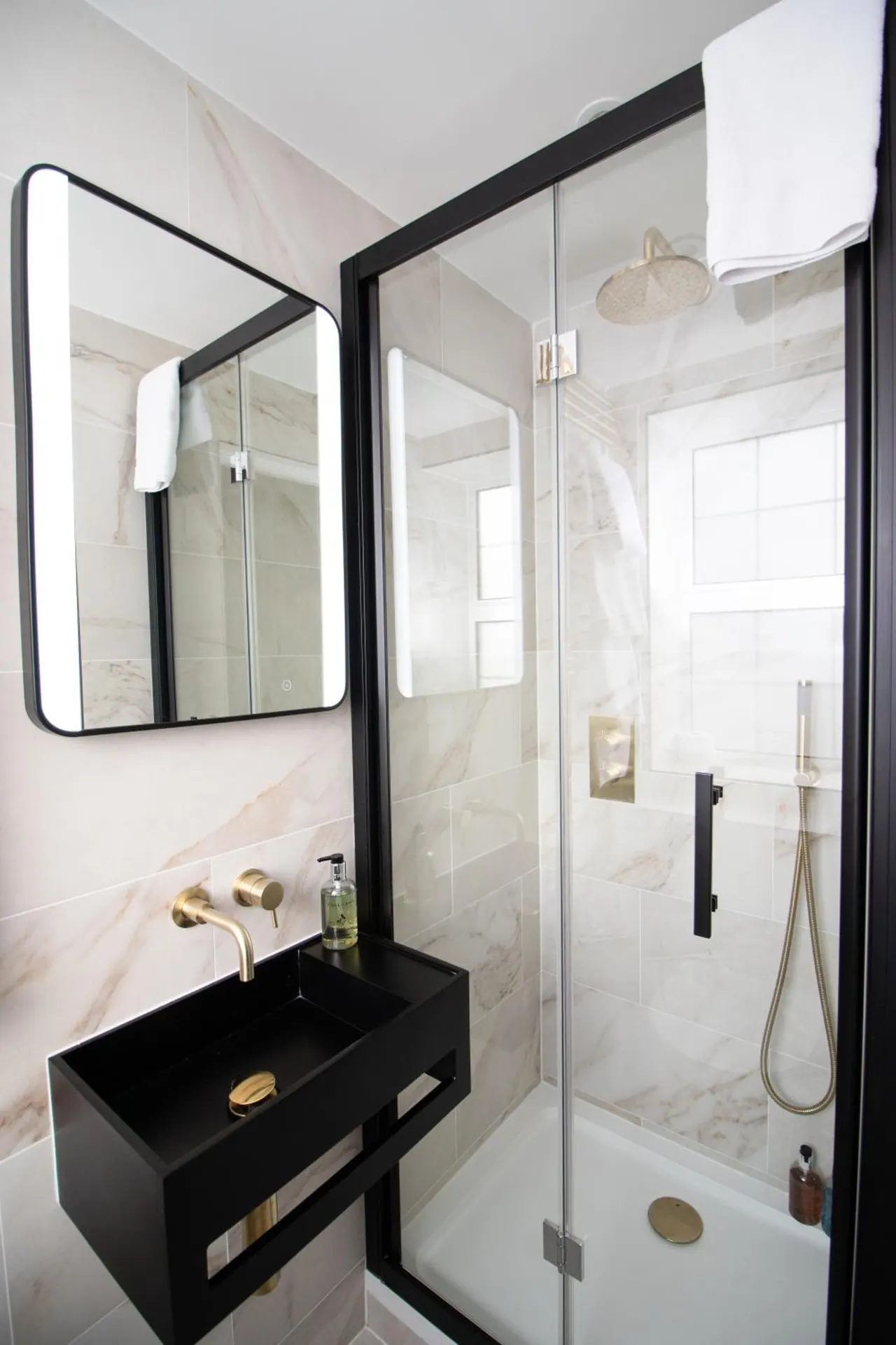 A small bathroom with a glass-enclosed walk-in shower, a black wall-hung basin with gold taps, and a backlit mirror.