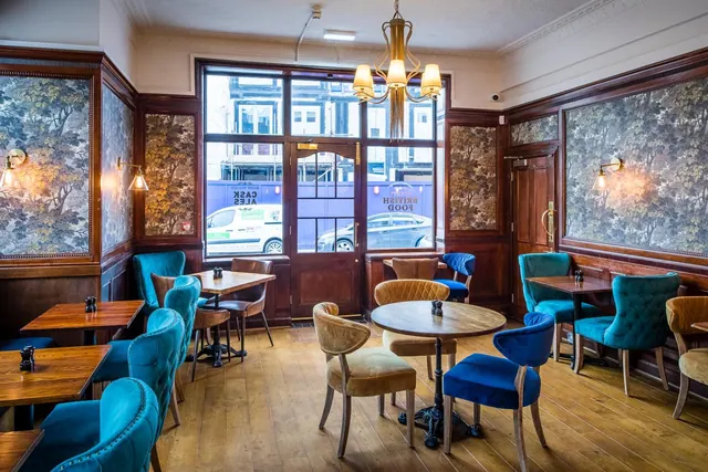 A dining room with teal and blue upholstered chairs, round wooden tables, wood-panelled walls and floral wallpaper, with a street view through the front window.