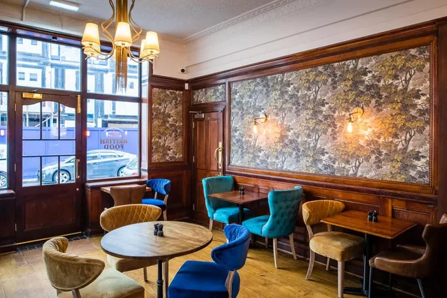 A restaurant dining area with velvet chairs, wooden tables and tree-patterned wallpaper, with a street visible through the front windows.