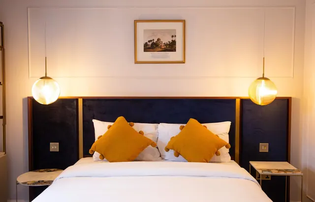 A double bed with a navy velvet headboard, mustard cushions and white bedding, flanked by two pendant lights and a framed print on the wall.