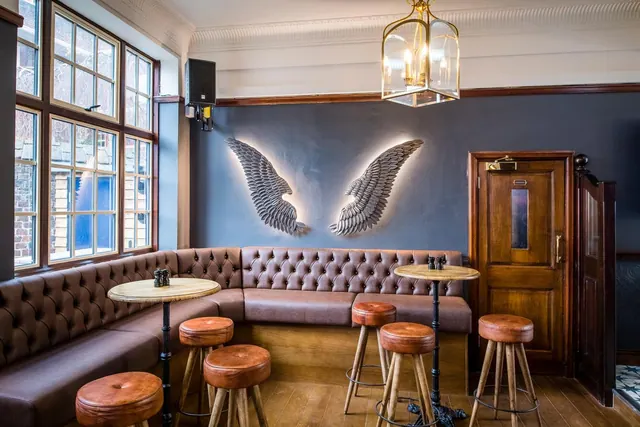 A lounge corner with a tufted leather banquette, round tables with leather stools, and a wall with two illuminated metal wing sculptures under a brass pendant lantern.