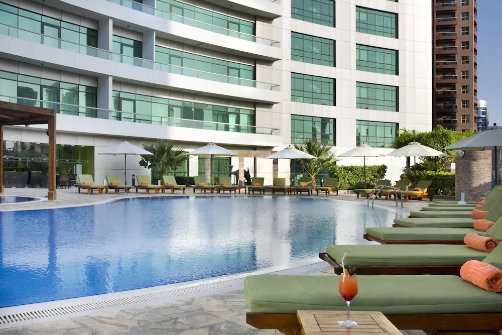 A large outdoor swimming pool with green sun loungers and parasols beside a multi-storey hotel building.