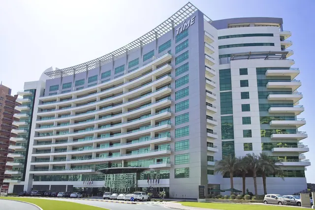 The exterior of a curved, multi-storey hotel building with the TIME sign on its facade, palm trees and parked cars at the entrance, under a clear sky.