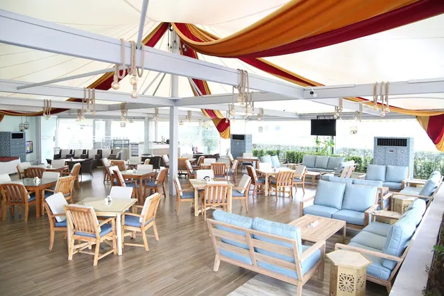 A covered terrace dining and lounge area with wooden tables, light blue sofas and chairs, and a draped fabric canopy, with hedges visible through the glass walls.