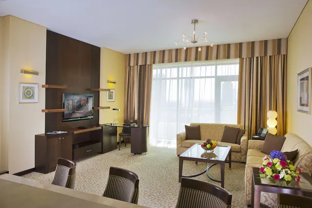 A hotel living room with two sofas, a wall-mounted television, a glass coffee table, a small glass desk and a large window with sheer curtains.