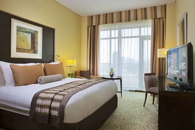 A hotel bedroom with a king bed, an armchair beside a round side table, a flat-screen television on a cabinet, and a window with sheer curtains looking out onto hazy distant buildings.