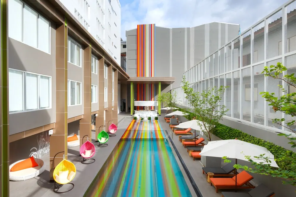 A long, narrow swimming pool with rainbow-striped tiles is surrounded by orange sun loungers under white umbrellas and hanging egg-shaped chairs.