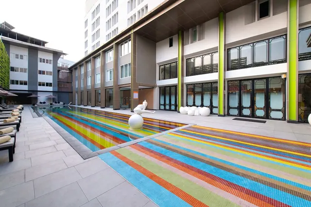A long pool with multicoloured striped mosaic tiles, white sculptures and sun loungers with towels beside it, in front of a multi-storey hotel building.