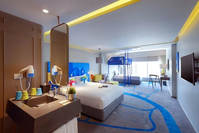 A hotel room with a king bed, a blue bunk bed with a ladder, a single bed by the window, a yellow seating bench, a desk and a wall-mounted television.