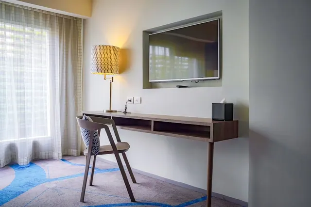 A hotel room corner with a wall-mounted television above a wooden desk shelf, a chair, a lamp and sheer curtains at the window.