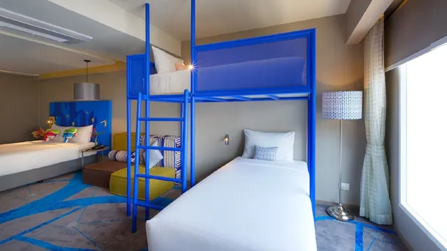 A bedroom with blue metal bunk beds, a double bed on the left, a striped seating area and a large window on the right.