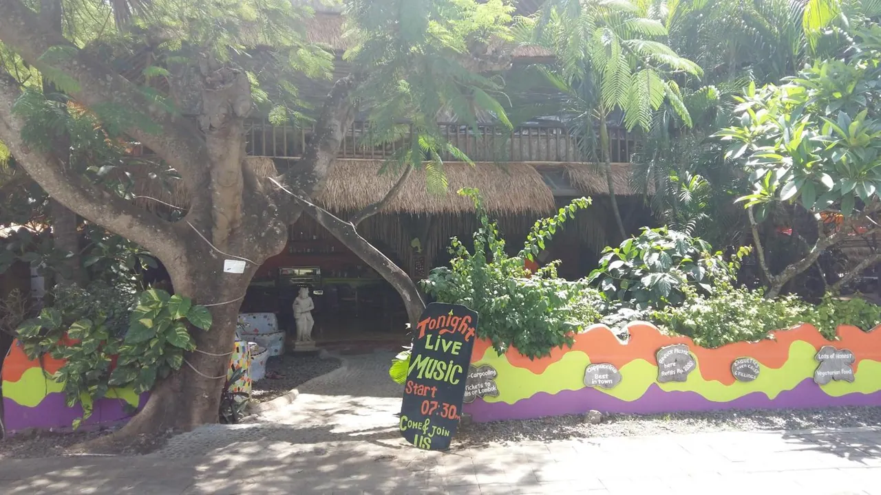 A thatched-roof building sits behind large trees, with a painted wall in front and a chalkboard sign advertising live music tonight at 07:30.