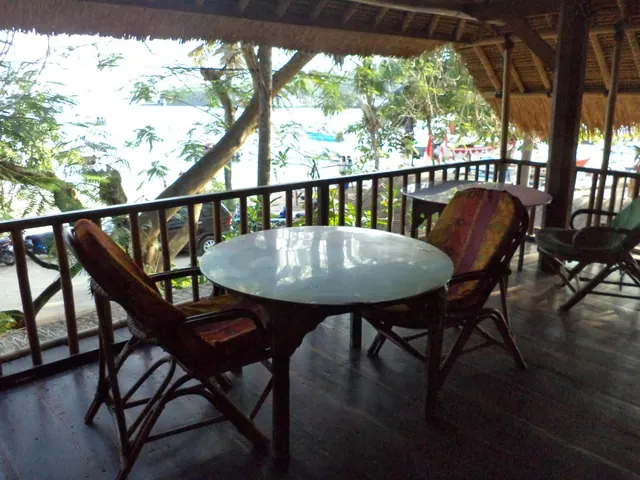 A covered thatched terrace with a round glass table and rattan chairs, overlooking trees and water with a parked car and boats beyond the railing.