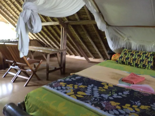 A bed with a mosquito net and colourful bedding sits beside wooden folding chairs and a table under a thatched roof.