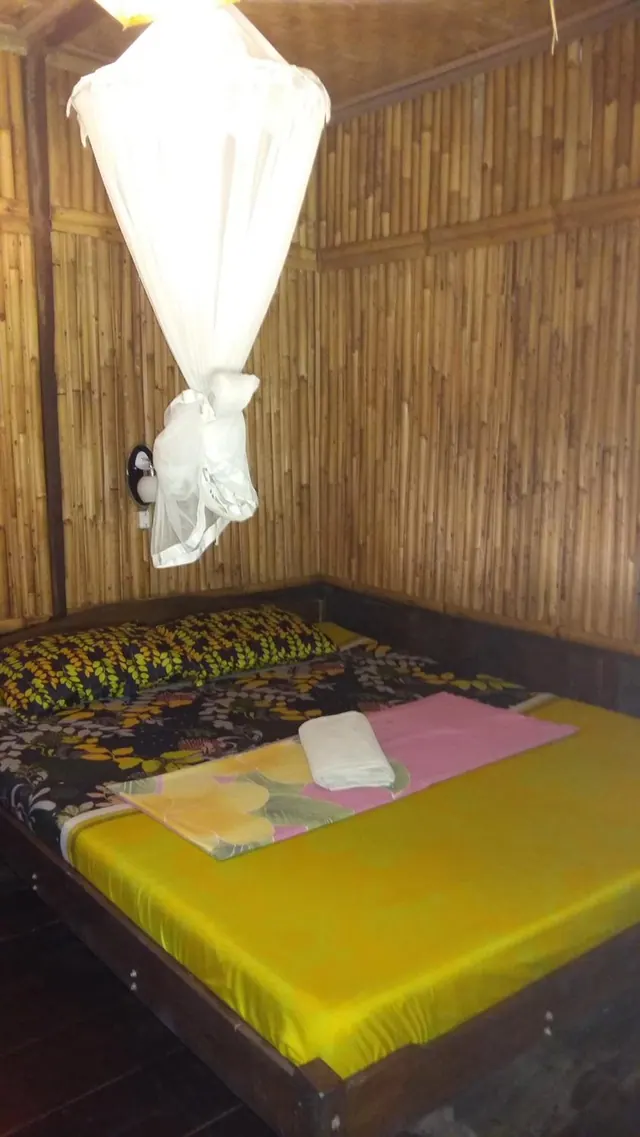A double bed with a yellow fitted sheet, floral bedding and a gathered white mosquito net sits in a bamboo-walled room.