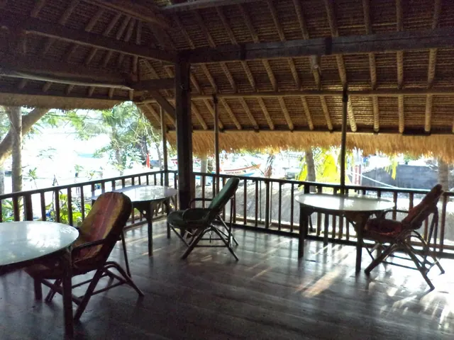 A thatched-roof open terrace with round tables and rattan chairs overlooks a beach with trees and moored boats.