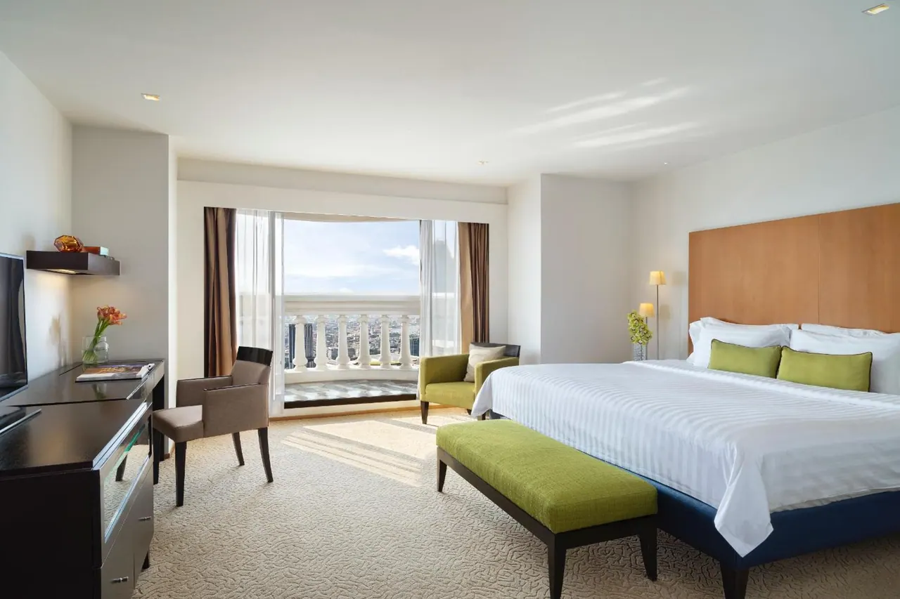 A bedroom with a king bed, green armchair and bench, a writing desk and television, with glass doors opening onto a balcony with a view of city buildings.