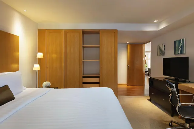 A bedroom with a large white bed, wooden wardrobe, bedside lamps, and a wall-mounted television above a desk with a chair.
