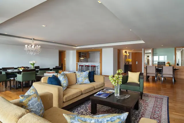A furnished living room with tan sofas, a dark wood coffee table on a patterned rug, and a dining area with a chandelier in the background.