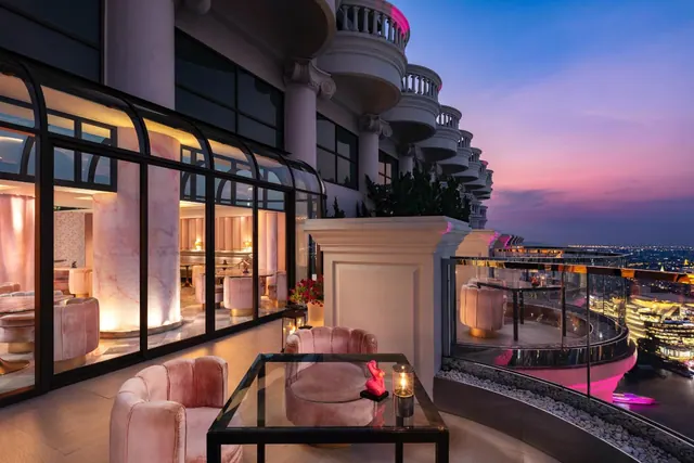 A terrace at dusk with pink velvet armchairs and a glass table, beside an illuminated interior lounge with a view over city lights.
