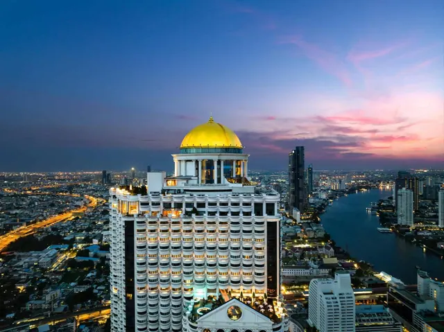 Aerial dusk view of a white high-rise hotel topped with a golden dome, with the city and a river visible behind it.