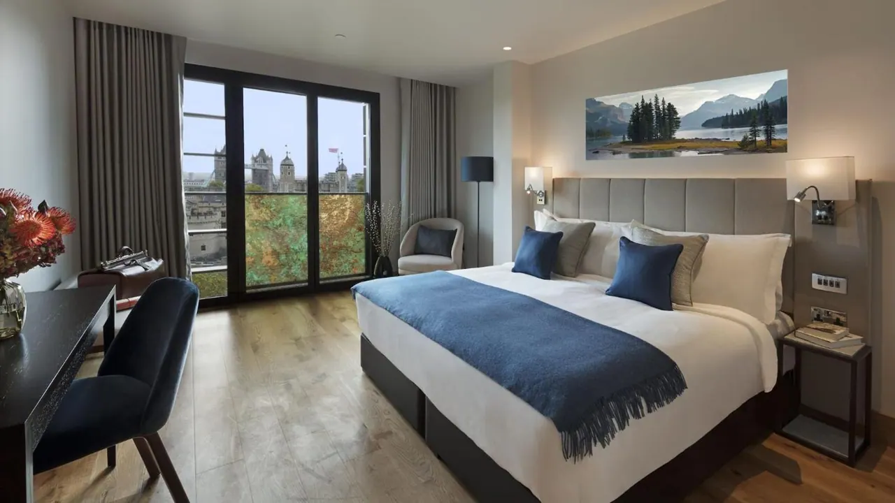 A bedroom with a king bed, blue throw and cushions, a desk and armchair, and floor-to-ceiling windows showing Tower Bridge outside.