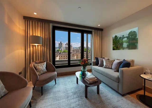 A living area with two armchairs, a grey sofa, a coffee table and a large window showing Tower Bridge and the city.