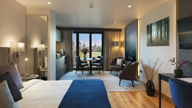 A hotel room with a king bed, a seating area with a round table and armchairs, and a large window looking out over city buildings.