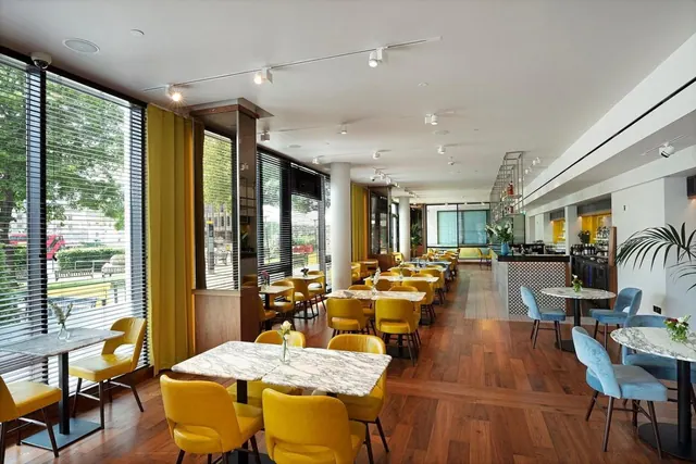 A dining room with yellow upholstered chairs and marble-topped tables, with floor-to-ceiling windows fitted with blinds that look out onto trees and a street.
