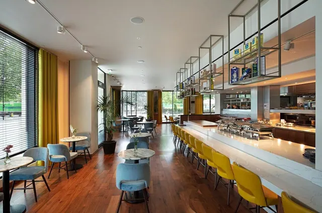 A restaurant dining room with blue and yellow chairs, round marble tables, and a buffet counter with chafing dishes beside tall windows.
