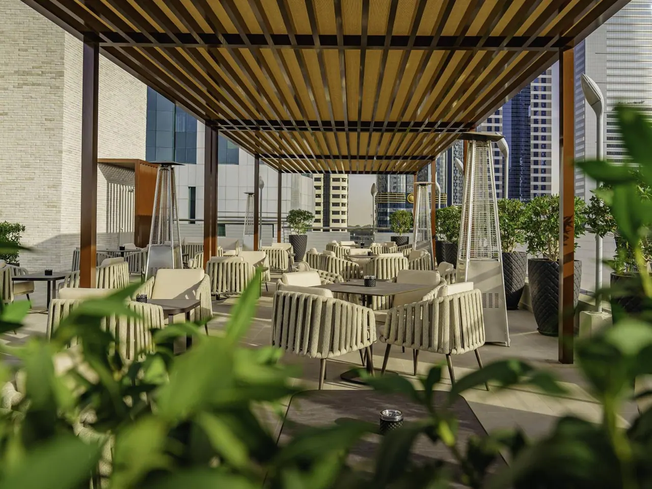 An outdoor terrace with cream rope-style chairs and tables under a wooden slatted pergola, with patio heaters, potted plants and high-rise towers behind.