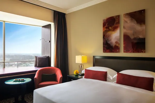 A bedroom with a large made bed, a red armchair and window seat, and a city view through a tall window.