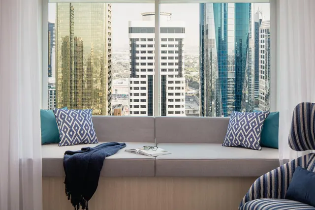 A window seat with cushions and a throw sits in front of a large window overlooking a cluster of high-rise towers.