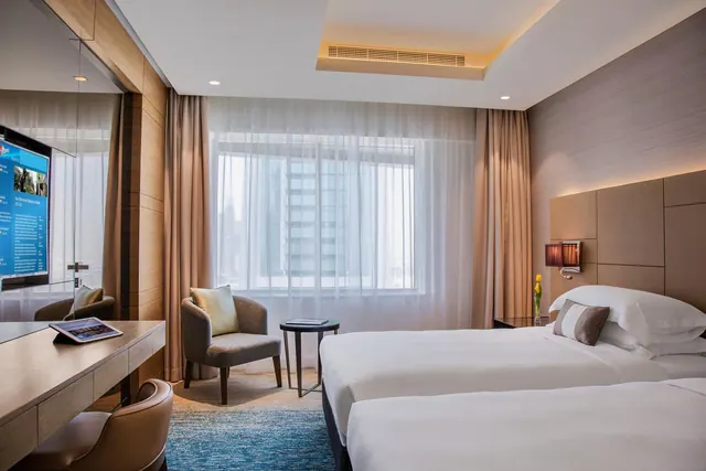 A hotel bedroom with two beds, an armchair and side table by a window overlooking high-rise buildings, and a desk with a wall-mounted television.