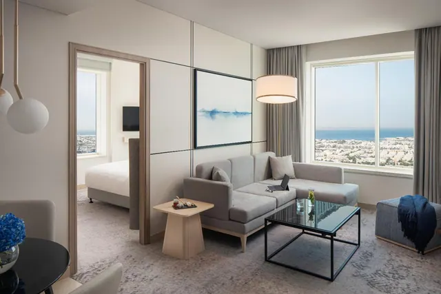 A living area with a grey sofa, a glass coffee table and large windows overlooking the sea and low-rise buildings, with a bed visible through the doorway.