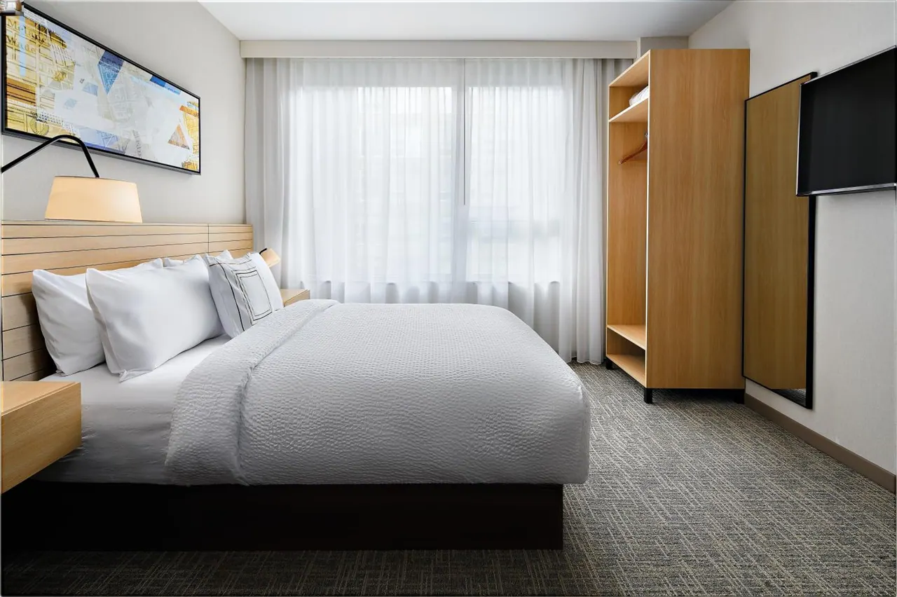 A bedroom with a king bed under white bedding, sheer window curtains, a wooden wardrobe and a wall-mounted television.