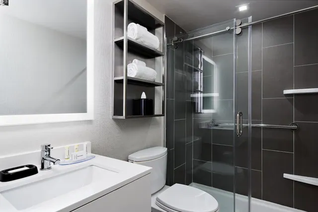 A bathroom with a single white vanity sink, a backlit mirror, open wall shelving holding folded towels, a toilet, and a glass-enclosed walk-in shower with dark tiles.