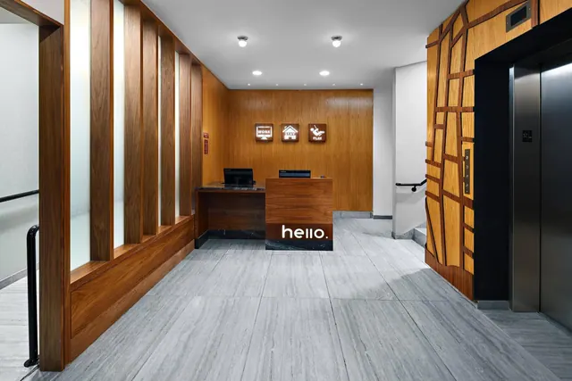 A hotel lobby with a wood-panelled reception desk bearing the word hello, wall signs reading WORK, STAY and PLAY, and a lift on the right.
