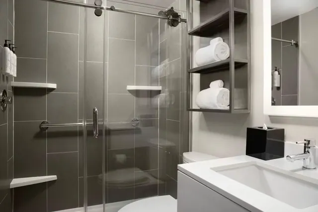 A walk-in shower with glass sliding doors and a grab rail sits beside a white vanity with a chrome faucet and rolled towels on open shelving.