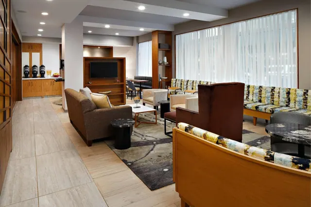 A lounge seating area in a hotel lobby has upholstered sofas and armchairs, a wall-mounted TV in a wood cabinet, and a beverage station at left beside tall sheer-curtained windows.
