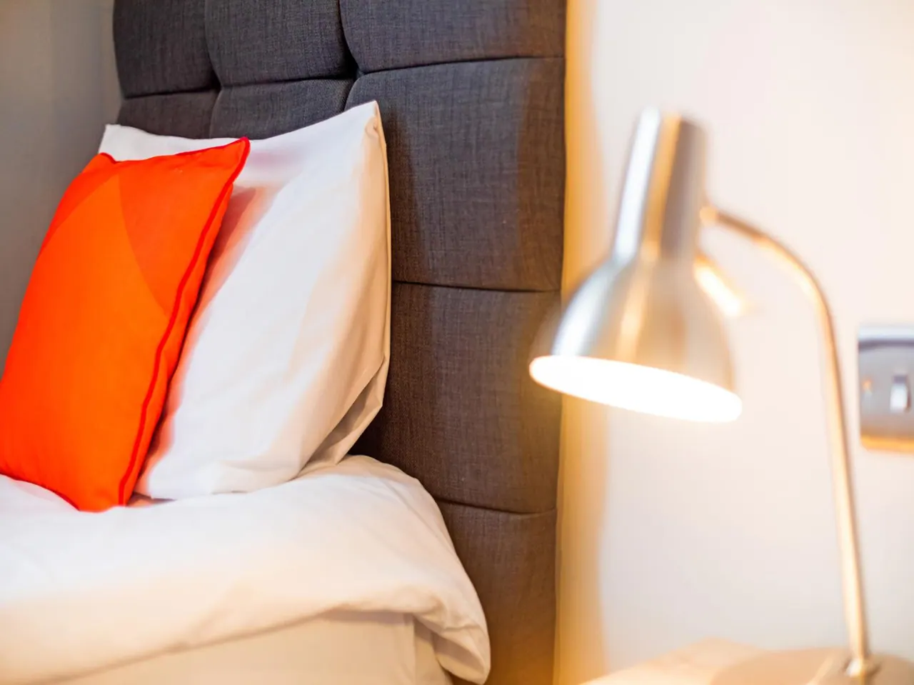 A made bed with white pillows, an orange cushion, a grey tufted headboard and a lit silver lamp beside it.