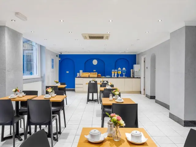 A breakfast dining room with wooden-topped tables, black chairs, a blue arched accent wall and a buffet counter with juice dispensers and bread baskets.