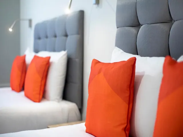 Two beds with grey tufted headboards, white bedding and orange cushions, with wall-mounted reading lights above them.